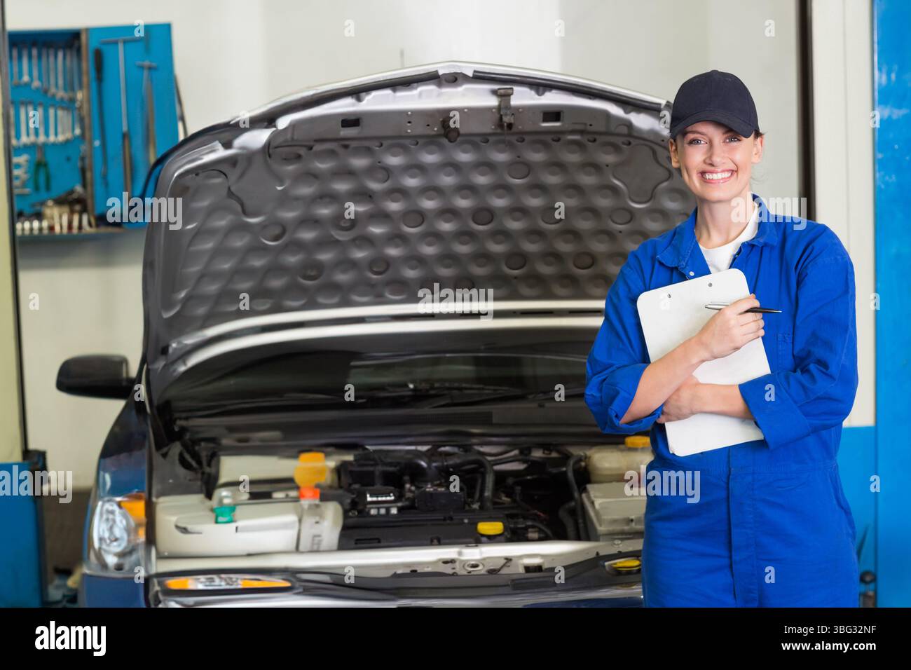 Mécanicien féminin en combinaison bleue vérifiant le capot de voiture ouvert dans l'atelier tenant le presse-papiers, espace copie Banque D'Images
