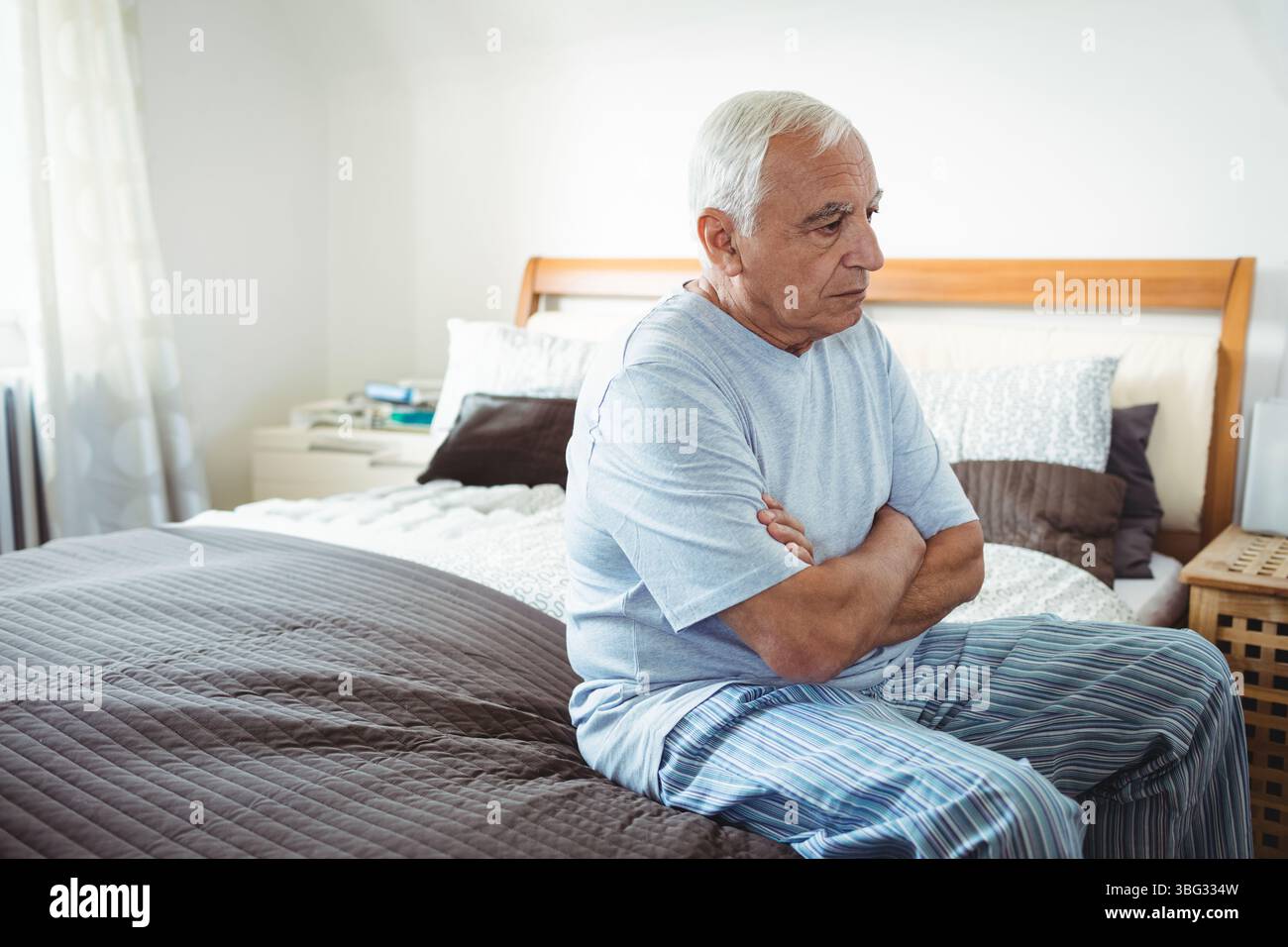 Homme aux cheveux blancs assis sur le bord du lit près de la table de nuit avec des livres, des oreillers, regardant directement dans la chambre Banque D'Images