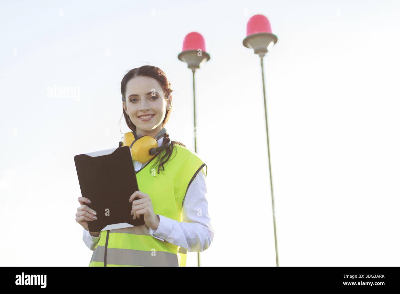 Ciel sur fond, jeune femme caucasienne brune dans l'ouest vert et les cache-oreilles jaunes se tiennent sur le toit, tiennent la tablette, regardent la caméra, montrent la tablette Banque D'Images