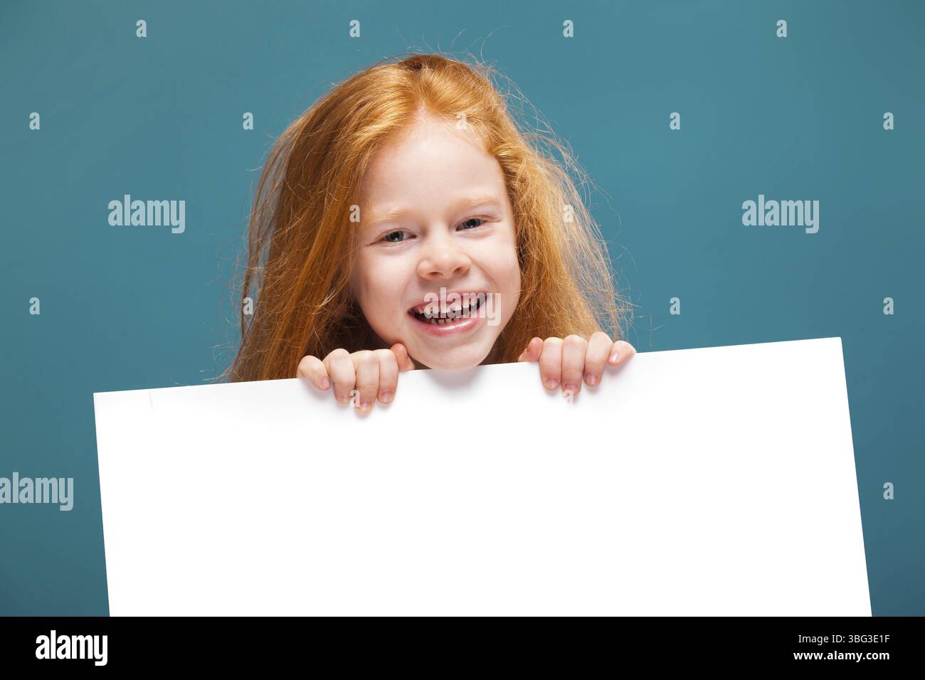 Isolé sur bleu, joli, caucasien rousse enfant en robe rayée bleue, avec de longs cheveux rouillés montrer une grande affiche vide, se cacher derrière l'affiche, souriant Banque D'Images