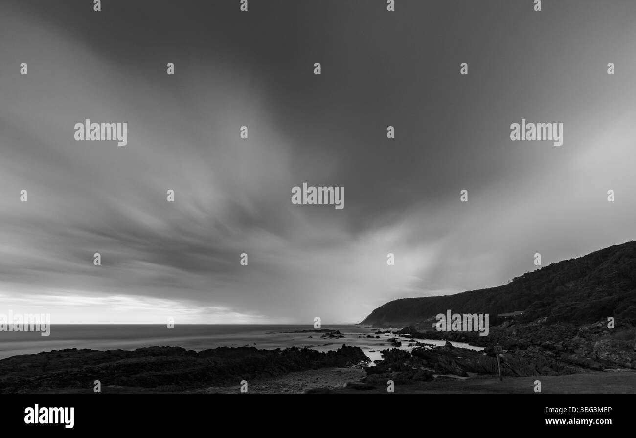 Longue exposition de nuages orageux sur une côte rocheuse près de Knysna, Afrique du Sud, avec des structures villageoises visibles au crépuscule. Banque D'Images