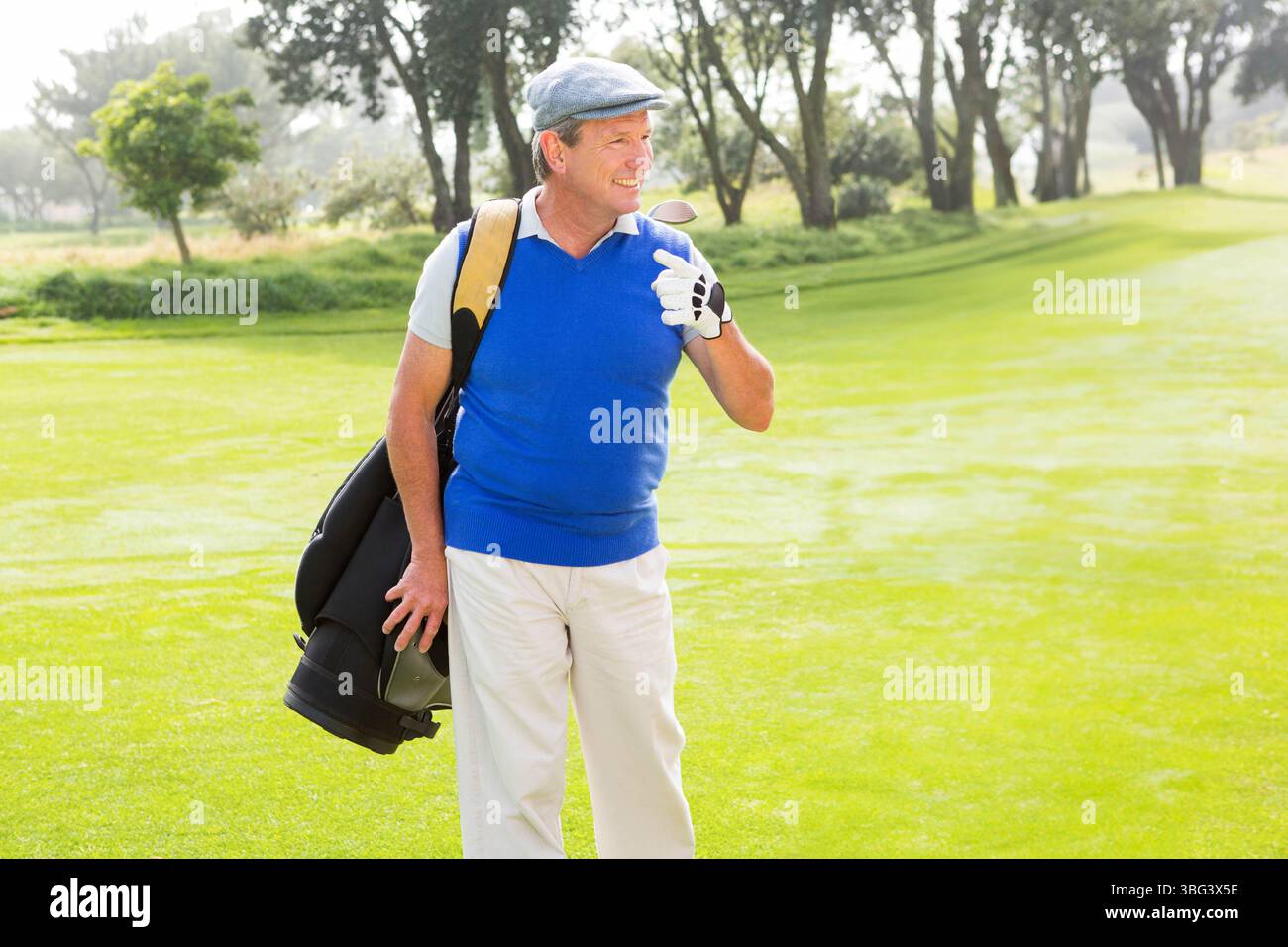 Homme senior souriant tout en portant un sac de golf, portant un gant de golf, casquette plate sur le fairway du parcours de golf Banque D'Images