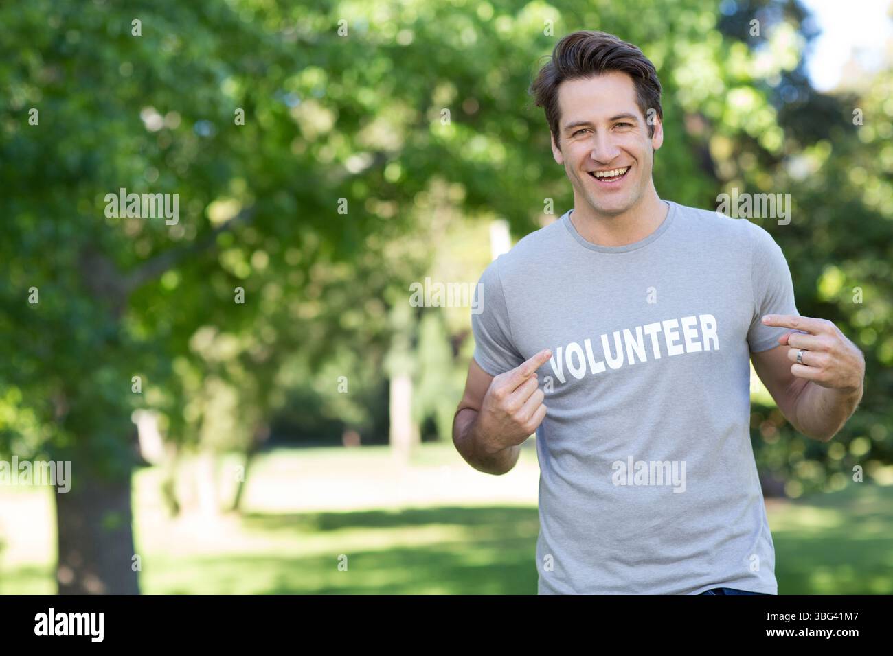 Volontaire masculin adulte debout dans un parc ensoleillé pointant le texte DU BÉNÉVOLE sur le t-shirt gris, espace de copie Banque D'Images