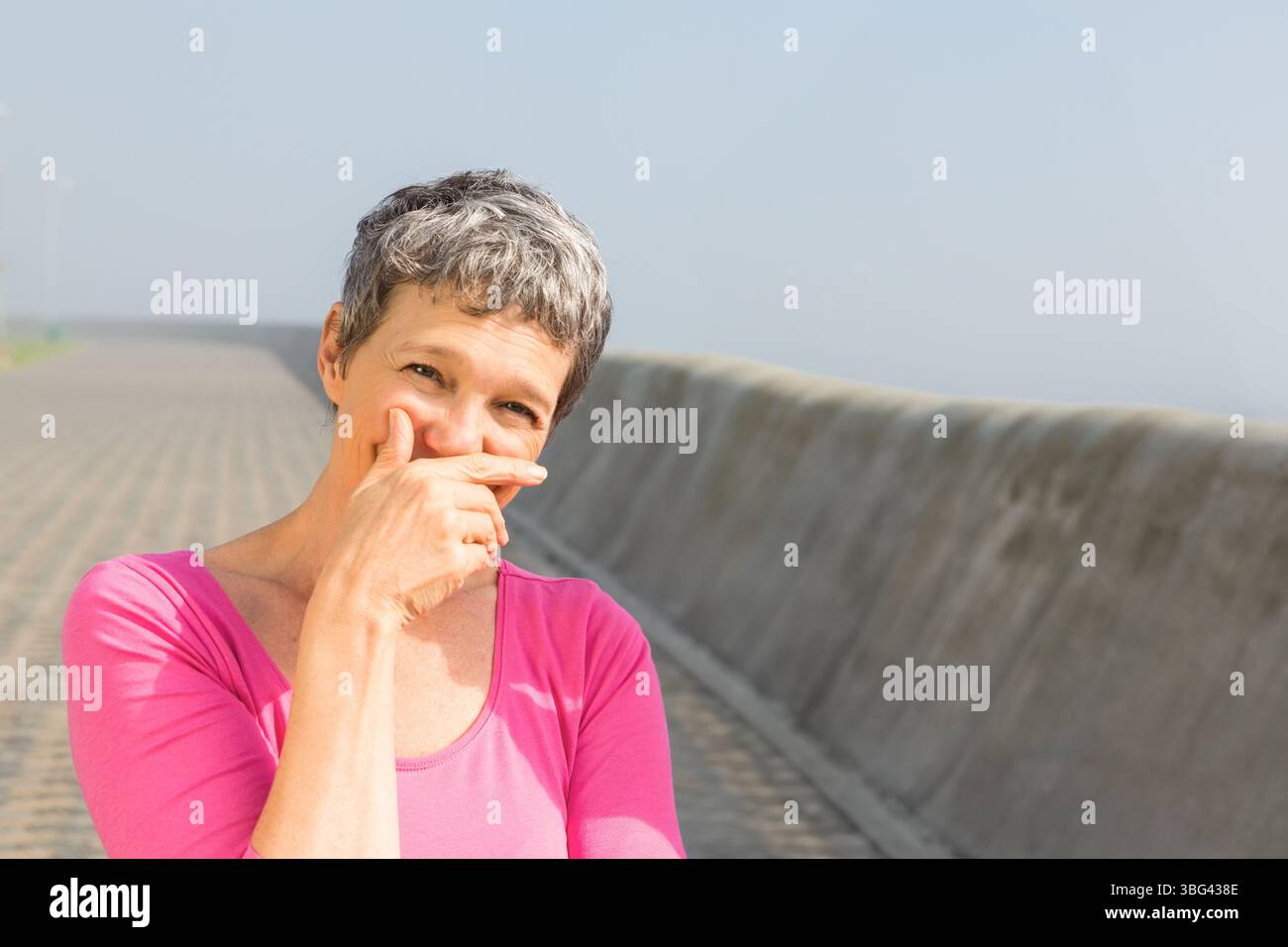 Femme senior portant le haut rose debout sur la passerelle faisant des gestes ludiques à côté de la barrière, copiez l'espace Banque D'Images