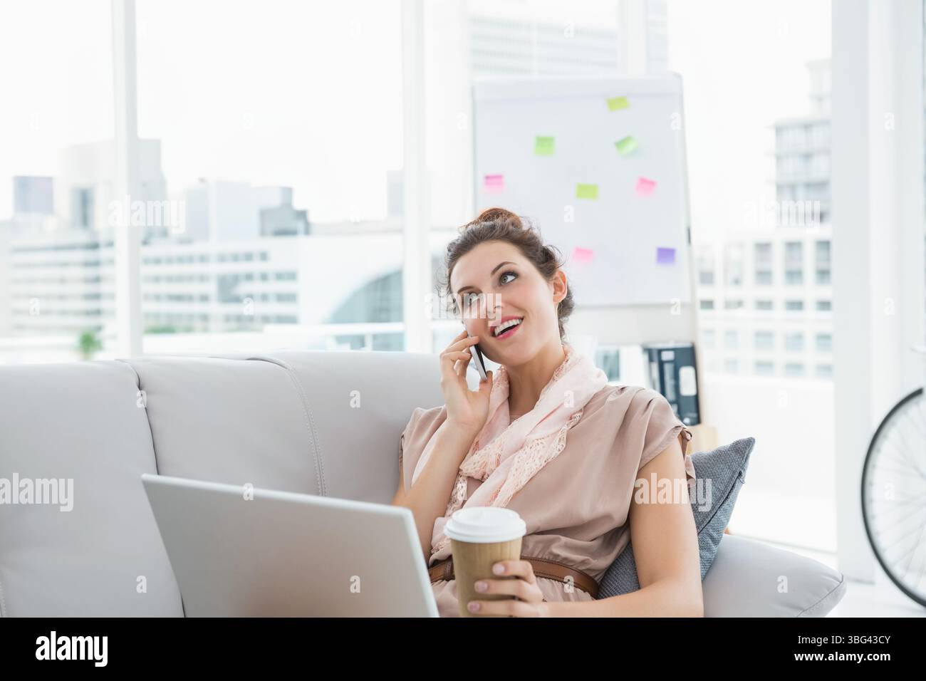 Femme d'affaires tenant smartphone à l'oreille avec tasse à café en utilisant l'ordinateur portable à côté de la roue de vélo au bureau Banque D'Images