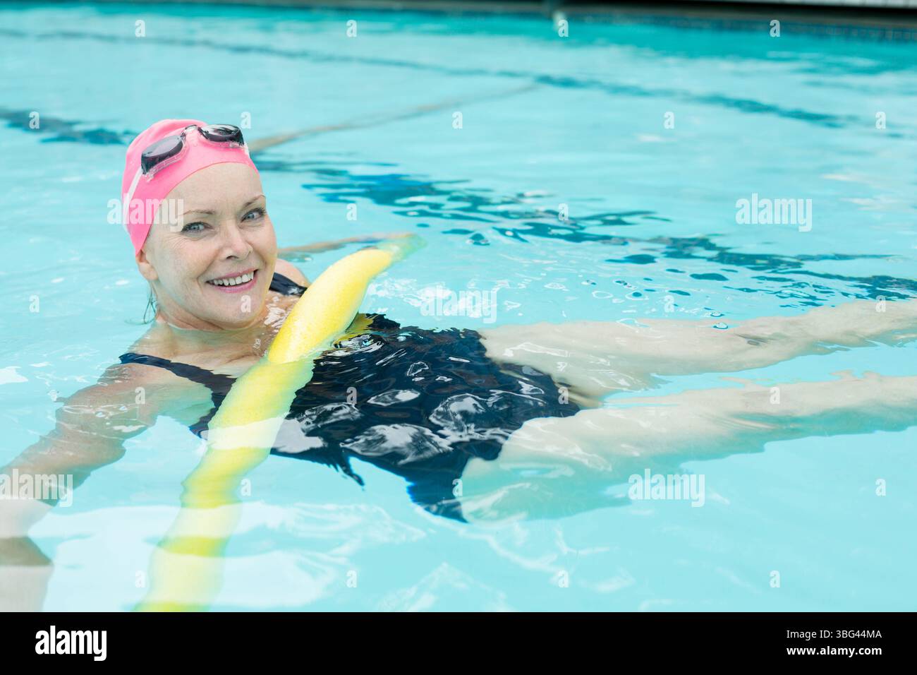 Femme senior flottant sur des nouilles jaunes à la piscine extérieure portant un maillot de bain noir bonnet de bain rose masques Banque D'Images