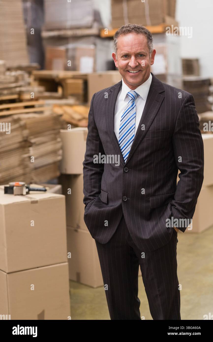 Homme d'âge moyen portant un costume d'affaires debout dans l'entrepôt parmi les boîtes, les palettes et le distributeur de ruban adhésif Banque D'Images