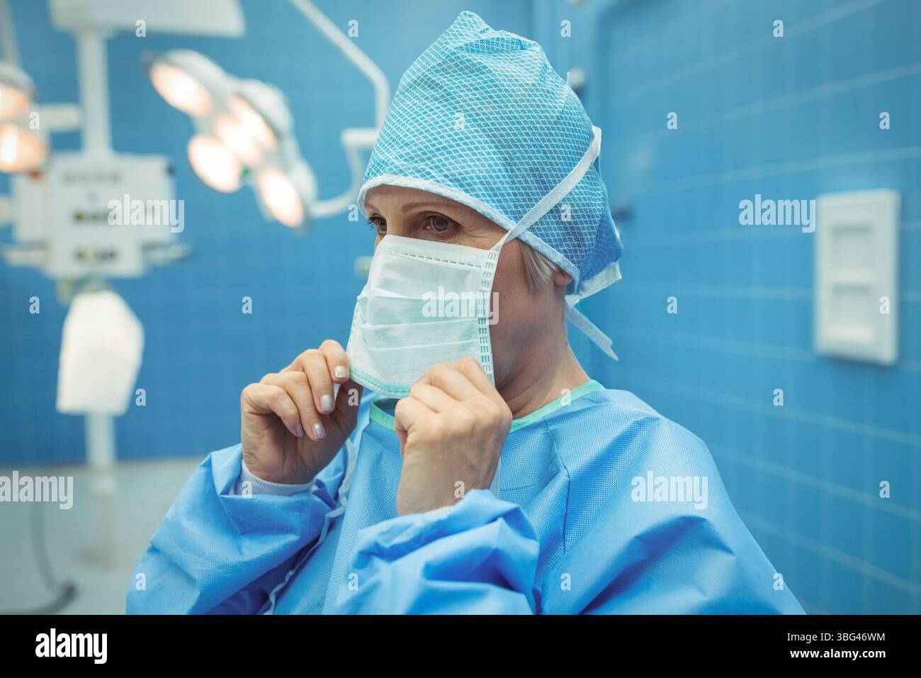 Chirurgien en gommage bleu ajustant le masque chirurgical dans OU avec les lumières chirurgicales et le panneau de commande Banque D'Images