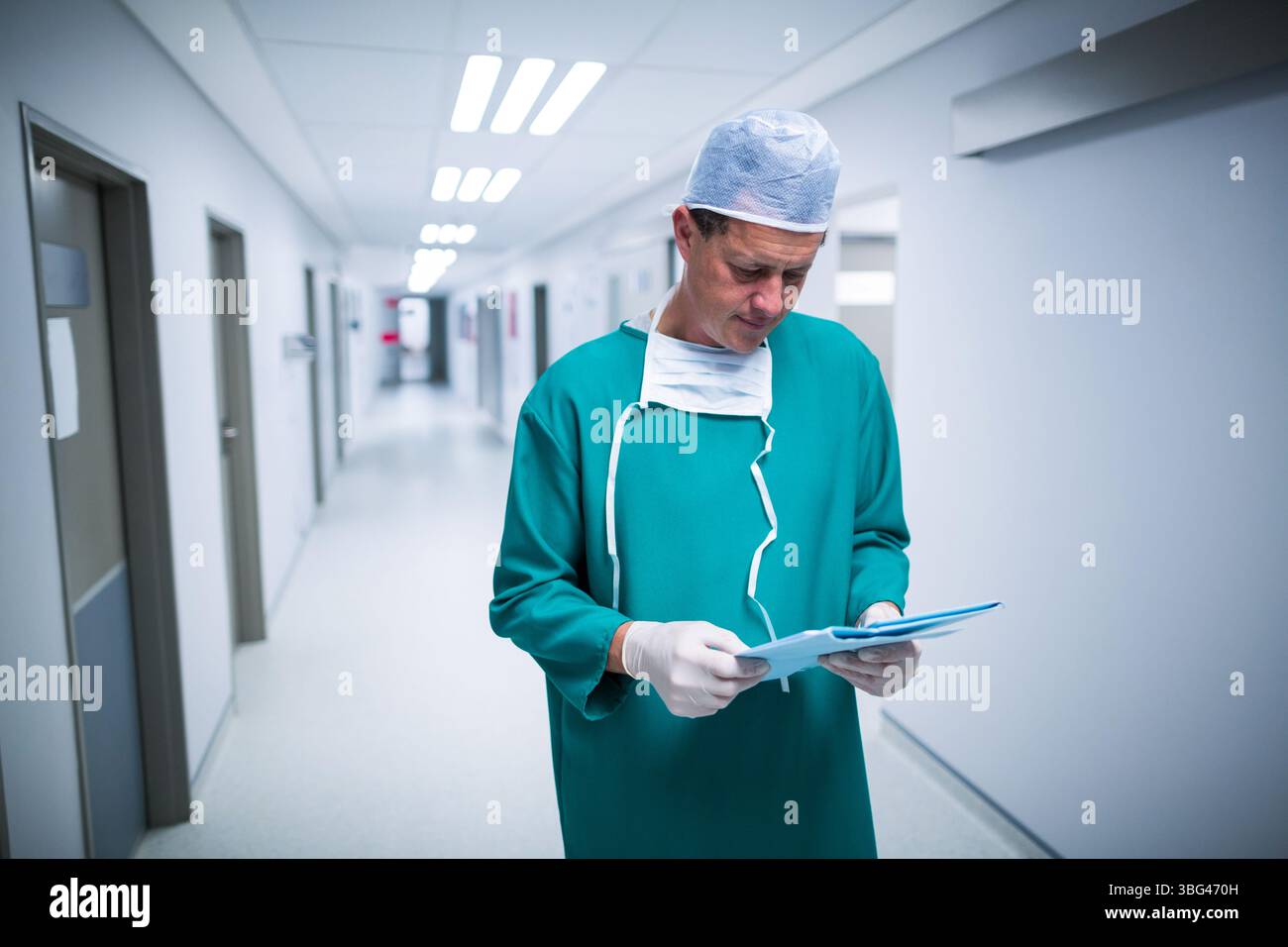 Homme adulte moyen debout lisant des cartes dans le couloir de l'hôpital portant des gommages, une casquette, un masque et des gants Banque D'Images