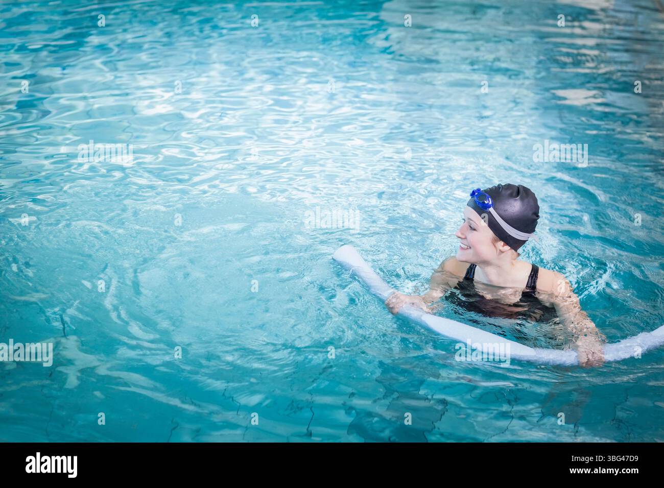Femme flottant avec des nouilles en mousse dans la piscine portant maillot de bain noir, bonnet de bain, lunettes bleues, espace de copie Banque D'Images