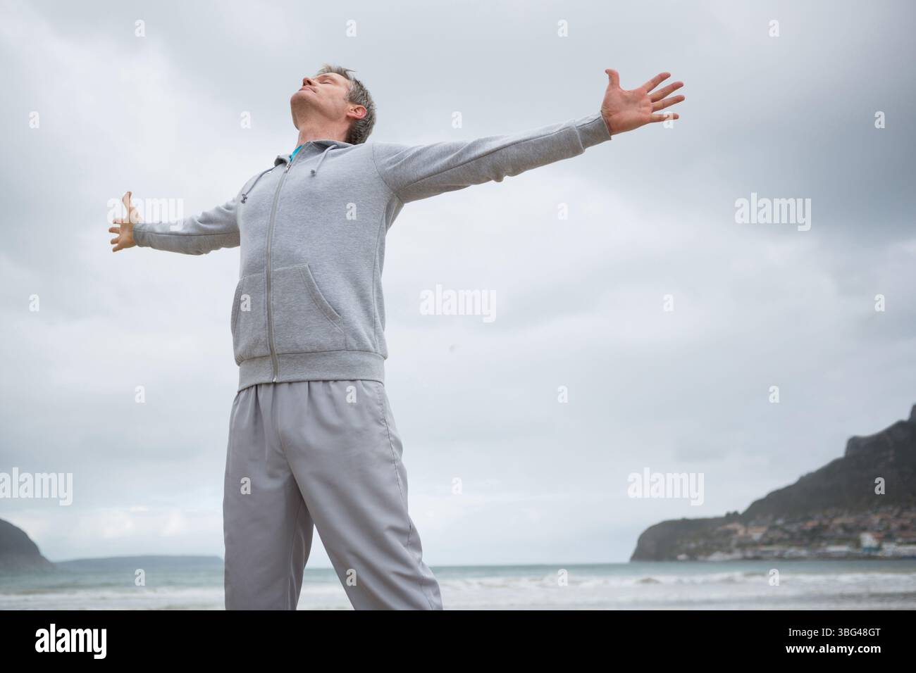 Homme d'âge moyen debout sur le rivage sablonneux levant les bras inclinant la tête vers l'arrière vers les vagues roulantes Banque D'Images