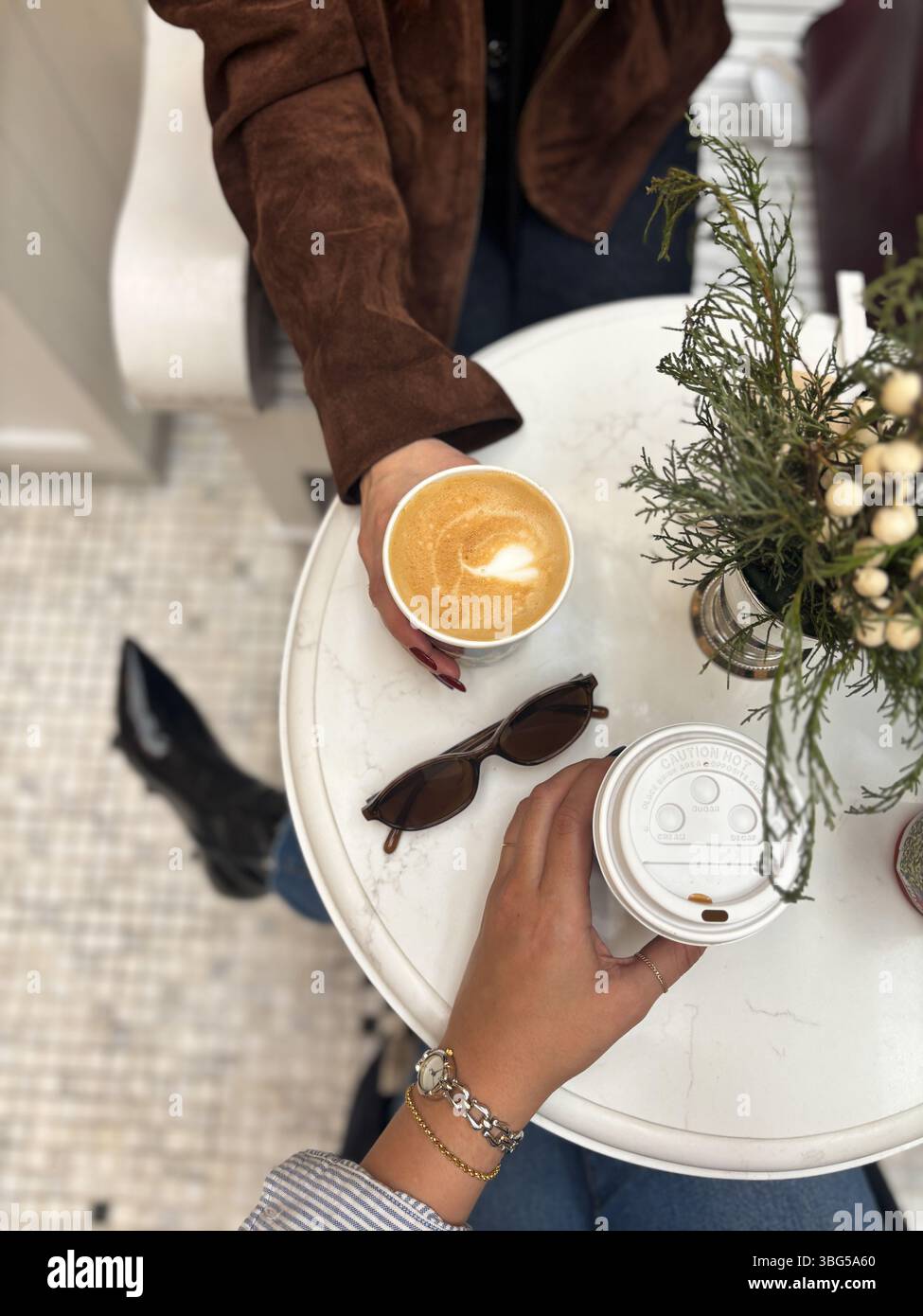 Mains tenant un latte avec des œuvres d'art en mousse en forme de coeur au-dessus d'une paire de lunettes de soleil et de verdure sur une table de café en marbre. Banque D'Images