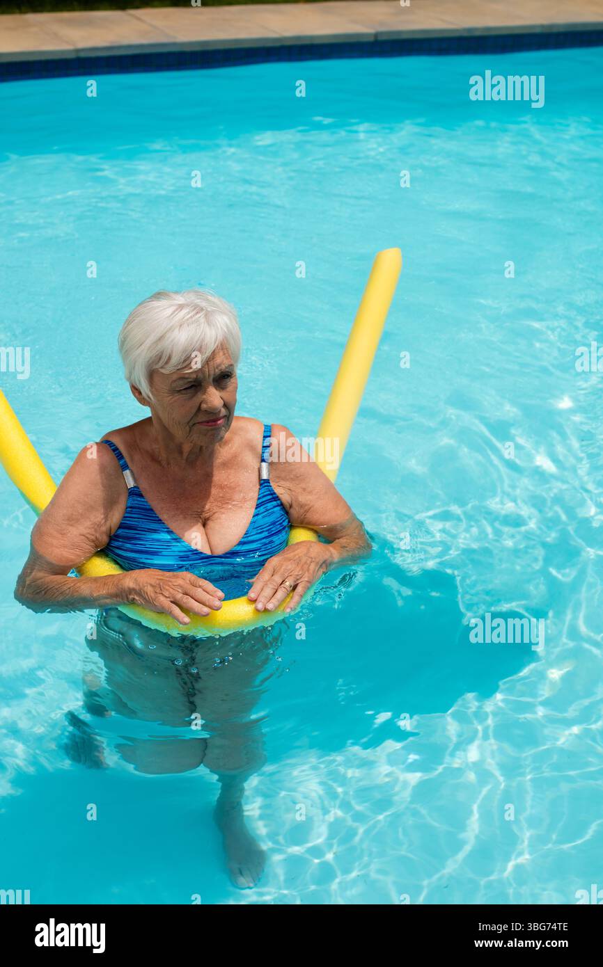 Femme senior pataugant dans la piscine de l'arrière-cour portant un maillot de bain à motifs bleus tenant des nouilles en mousse jaune Banque D'Images