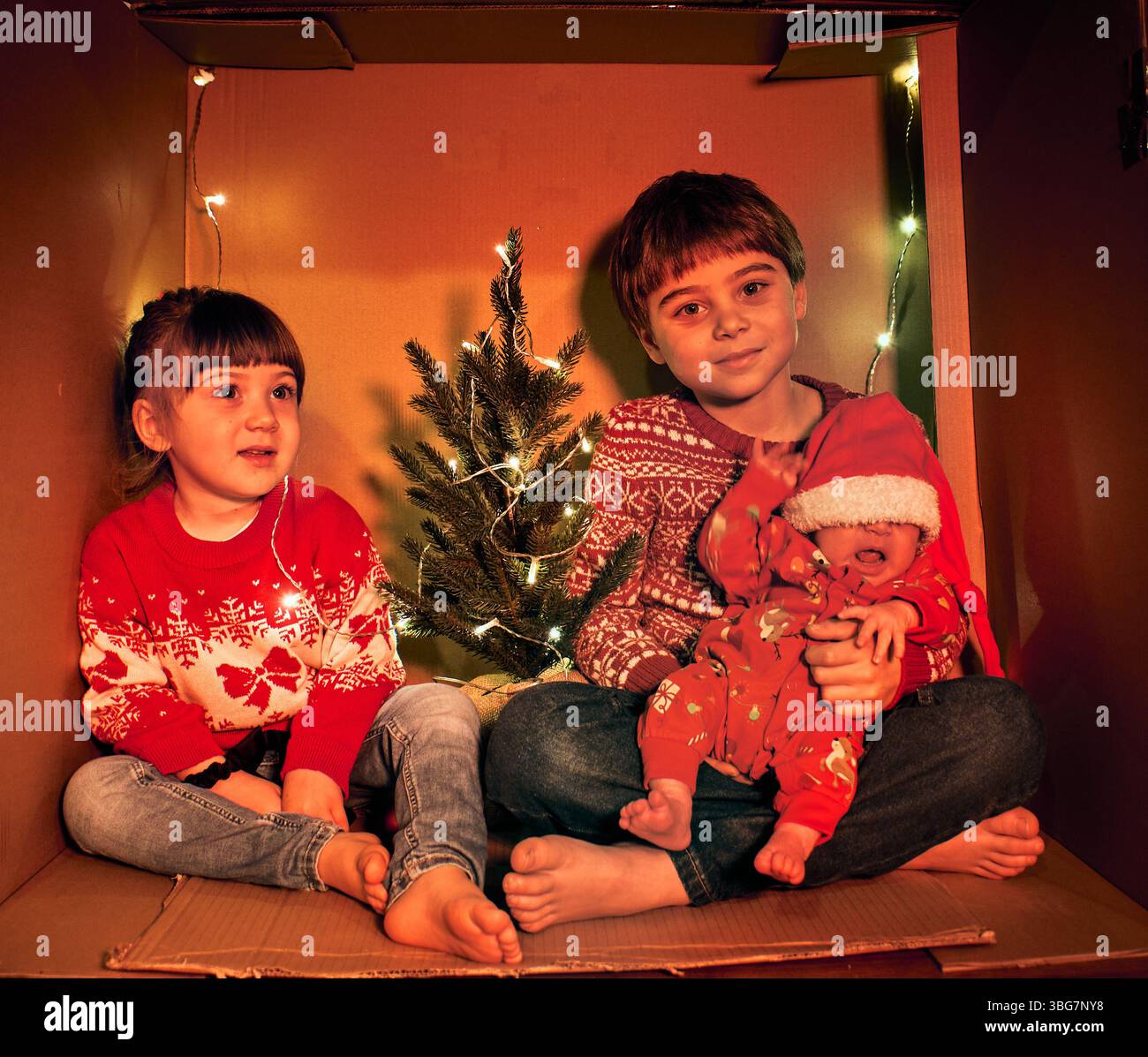 Trois enfants sont assis dans une boîte en carton confortable décorée de lumières de fée et d'un petit arbre. Vêtus de pulls festifs, ils créent un Noël chaleureux et bricoleur Banque D'Images