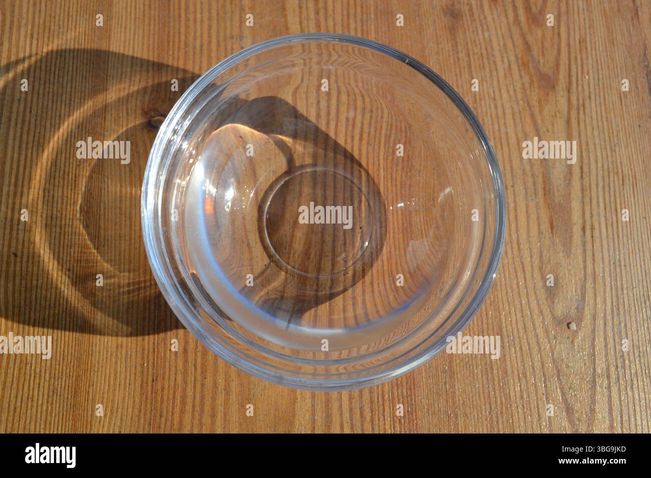 Vue de dessus du bol transparent en verre vide comme une tasse sous la lumière du soleil avec son ombre sur fond de table en bois rayé brun. Restauration à boire. Banque D'Images