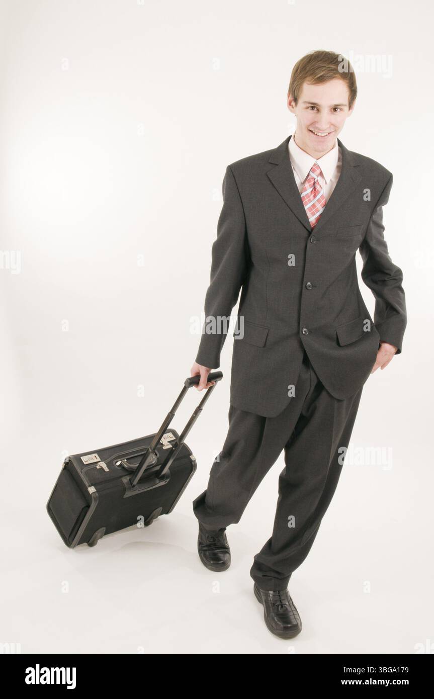 Photo du corps entier d'un jeune homme en costume sombre marchant vers la caméra, tirant la valise d'un pilote derrière lui, souriant à nouveau dans la caméra Banque D'Images