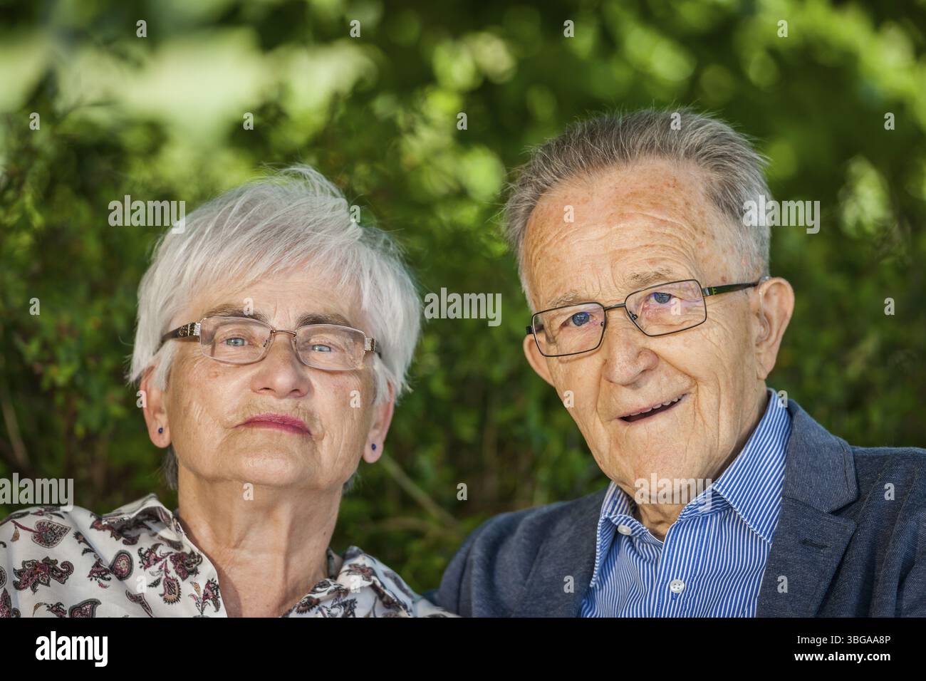 Portrait de la tête et des épaules d'un couple de retraités âgés avec des lunettes devant des feuilles vertes en été regardant joyeusement dans la caméra Banque D'Images