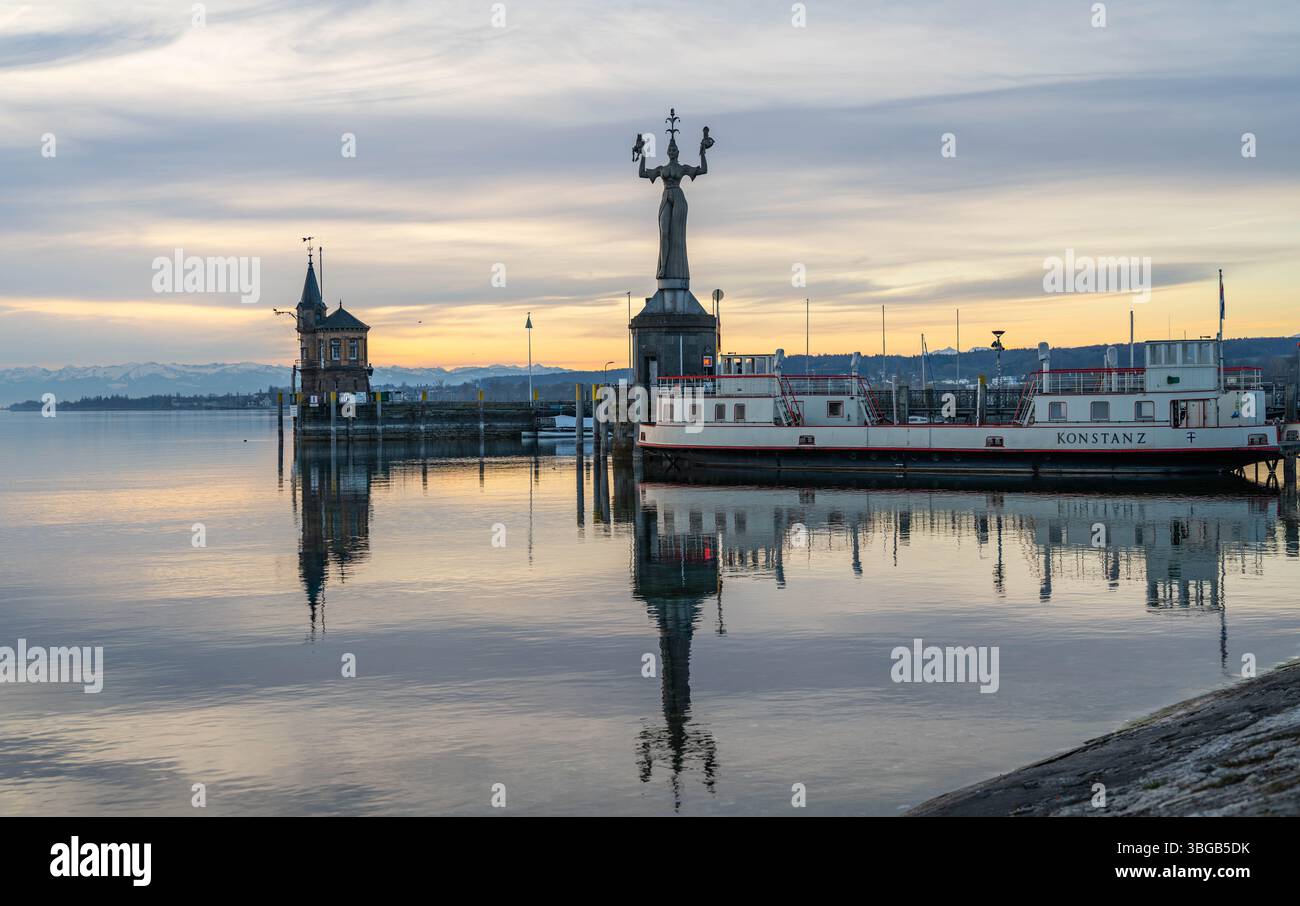 Beschreibung : Die Imperia-Statue, der Leuchtturm und die traditionelle Autofähre an der Hafeneinfahrt mit dem ruhigen See und den Alpen am Horizont in Banque D'Images