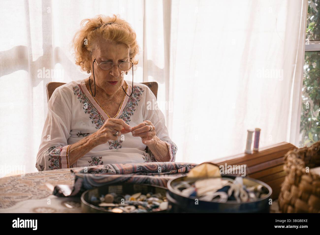 Couturière sénior concentrée enfilant méticuleusement une aiguille, entourée de matériaux à coudre sur une table à la maison, éclairée par la lumière naturelle Banque D'Images