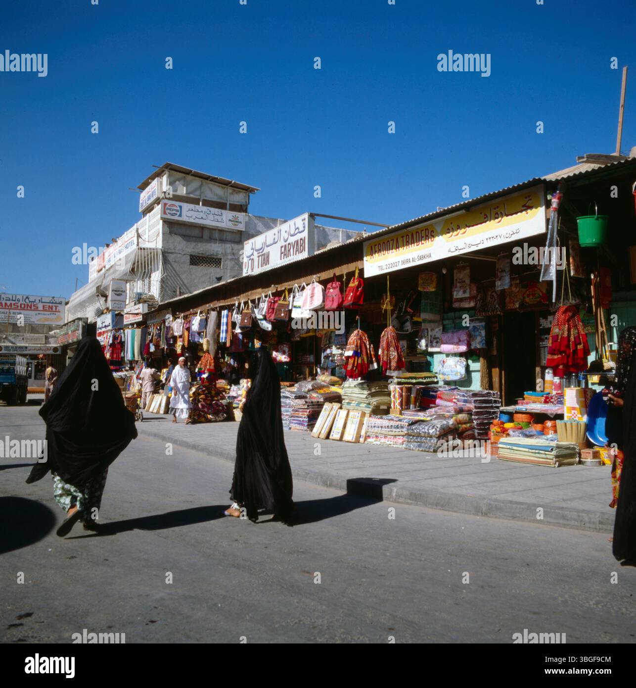 Straßenabschnitt mit kleinen Einzelhandelsgeschäften und Warenauslagen im Freien in einem Marktviertel von Dubai. Zwei Frauen in schwarzer Kleidung gehen auf dem Gehweg an den Läden vorbei. Banque D'Images