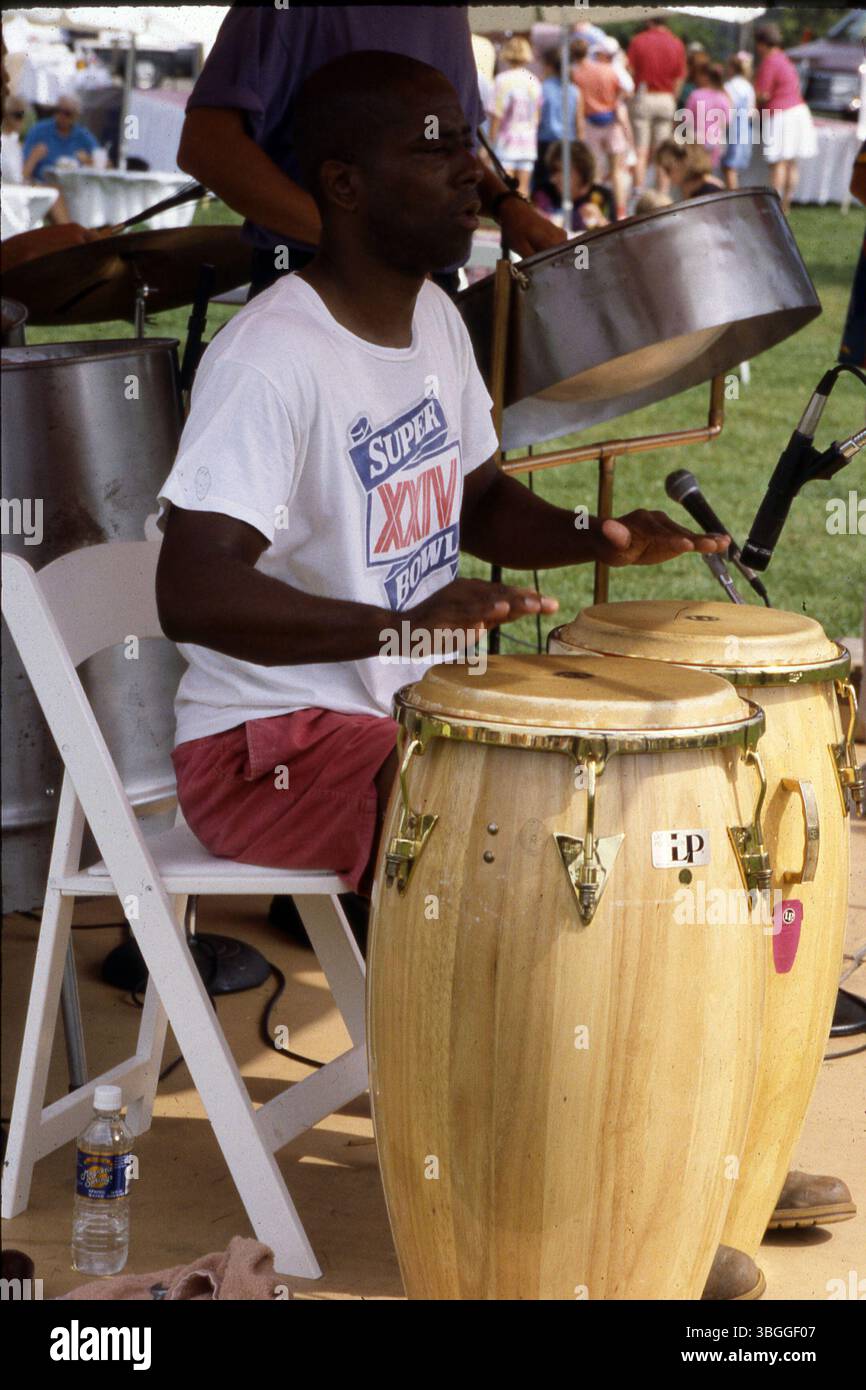 En juillet 1980, des musiciens se produisent avec des bongos et des tambours en acier lors d'un événement en plein air. Le joueur de Bongo porte un t-shirt Superbowl XXIV et un short rouge, tandis qu'un microphone capte le son. Les visiteurs sont assis à des tables près de kiosques couverts, créant une atmosphère animée et interactive. Banque D'Images