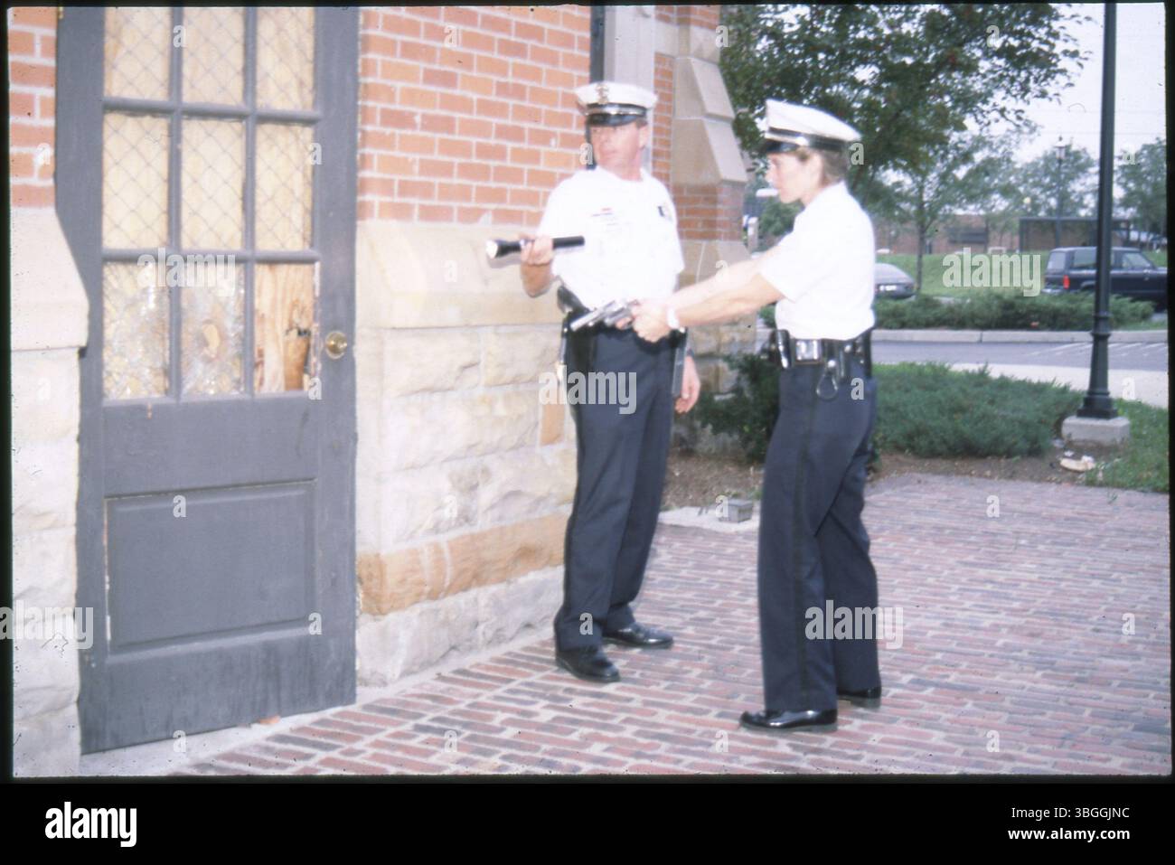 Deux policiers de Columbus sont vus à l'extérieur d'un bâtiment qui semble avoir été cambriolé. L'officier masculin tient une lampe de poche dans sa main droite, tandis que l'officier féminin a son arme tirée. L'image a été prise vers 1980. Banque D'Images