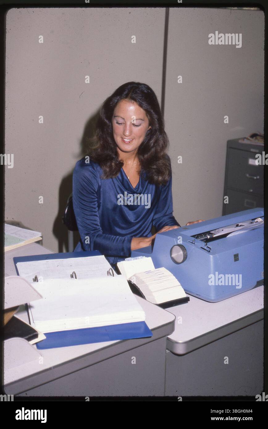 Une employée de bureau non identifiée utilise une machine à écrire, avec un rolodex et des classeurs métalliques visibles sur le bureau. La photo a été prise vers 1971-1980. Banque D'Images