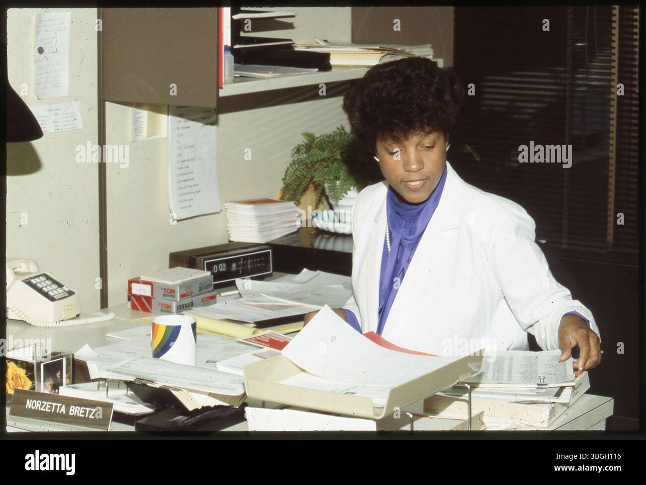 Norzetta Bretz, employée de bureau, est représentée assise à son bureau, entourée de piles de papier. Un téléphone et un réveil/radio sont visibles sur le bureau. Cette image date des années 1970 ou 1980 et reflète un environnement de bureau typique de cette époque. Banque D'Images