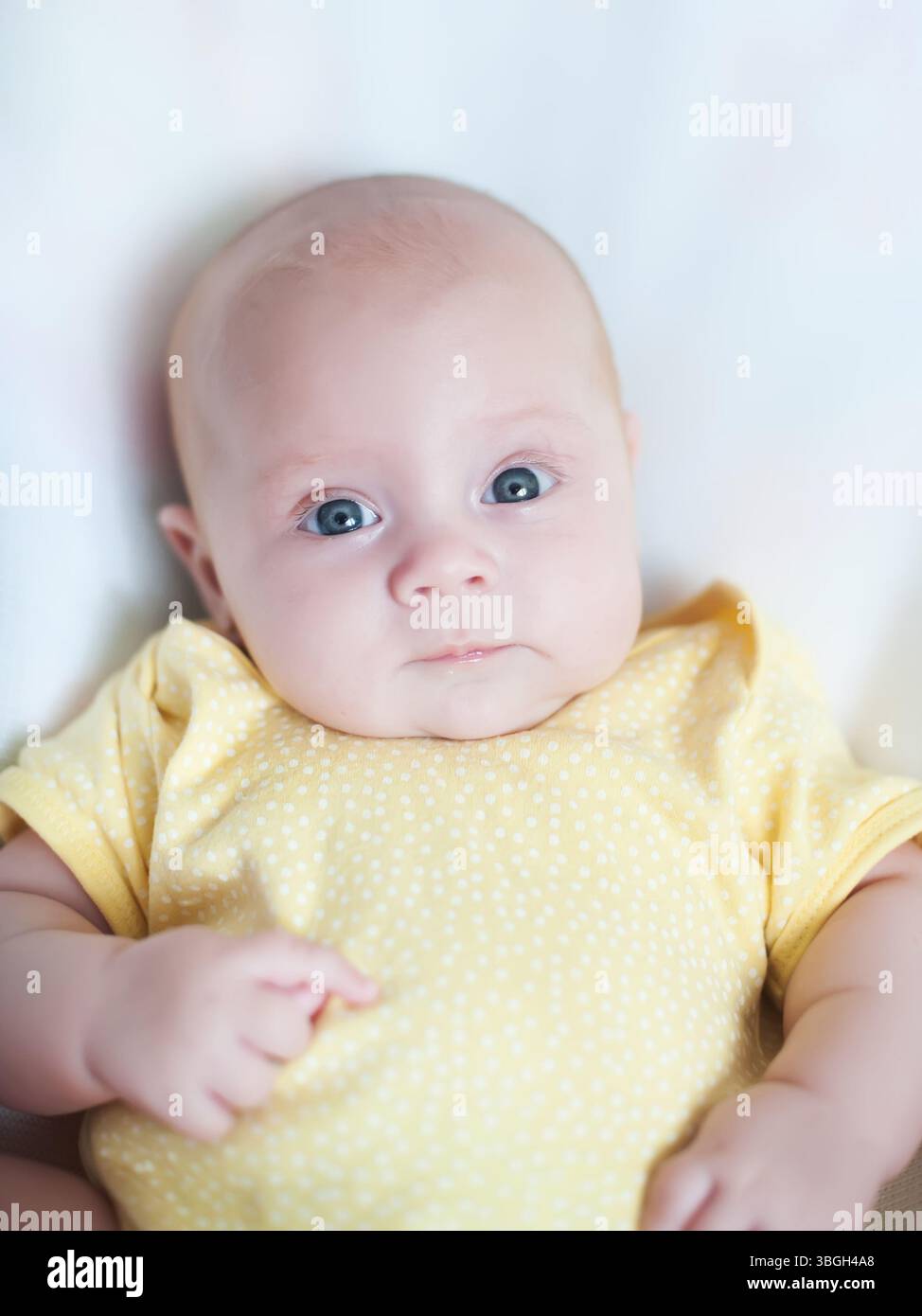 Bébé aux yeux bleus est habillé dans une combinaison jaune à pois, couché sur une surface douce, affichant une expression curieuse et innocente dans un environnement serein Banque D'Images