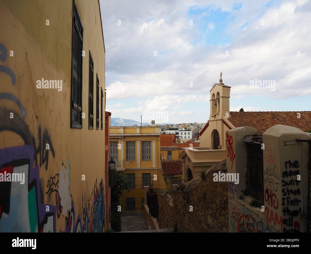 Rue calme dans le quartier de Plaka, Athènes, avec des murs jaunes recouverts de graffitis, une petite église et des paysages authentiques sous un ciel nuageux. Banque D'Images