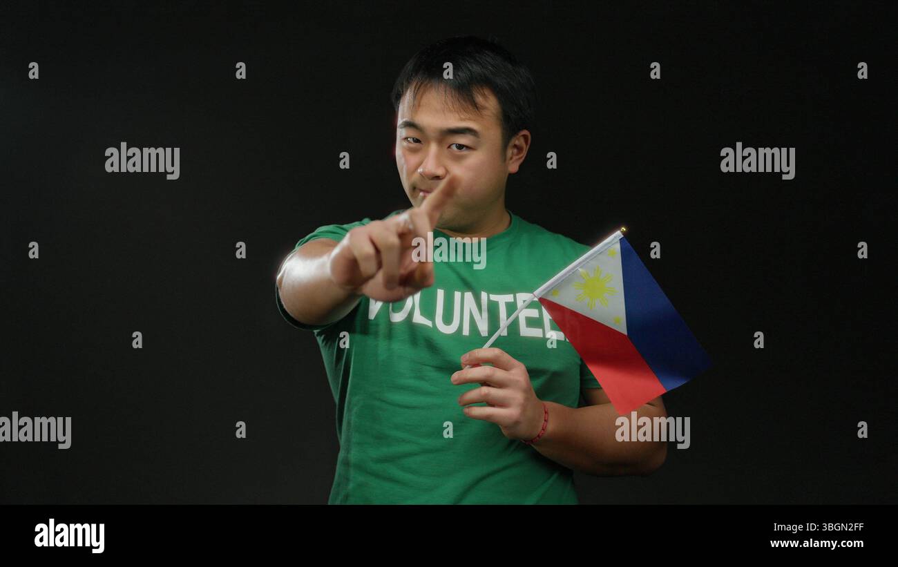 Jeune homme en chemise verte tenant le drapeau philippin sur fond noir faisant des gestes de la main, représentant le bénévolat et la fierté nationale. Banque D'Images