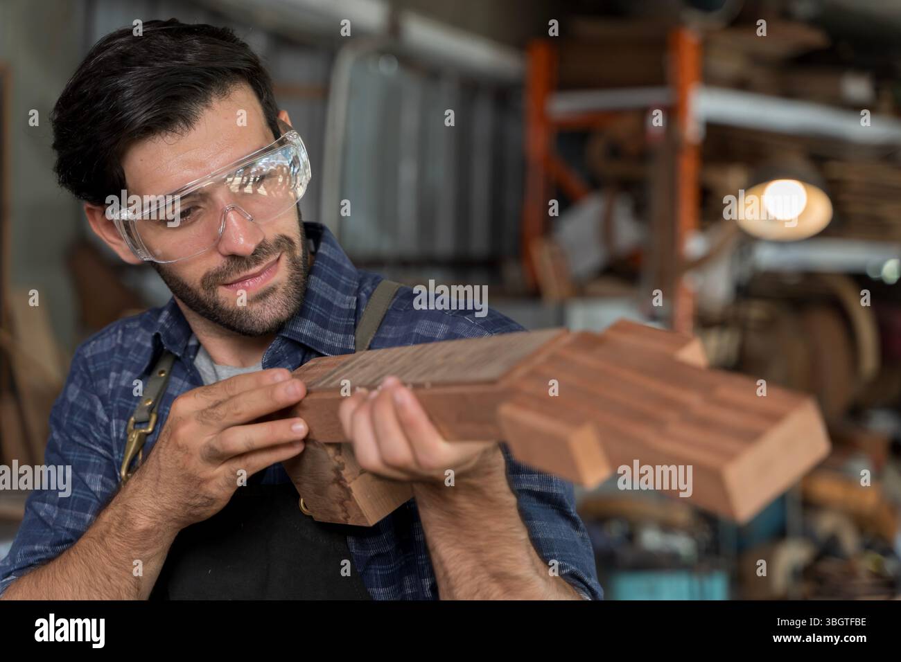 Portrait d'un charpentier masculin qualifié examinant soigneusement un cou de guitare en bois dans un atelier. Porter des lunettes de sécurité pour la protection, mettant en vedette artisan Banque D'Images