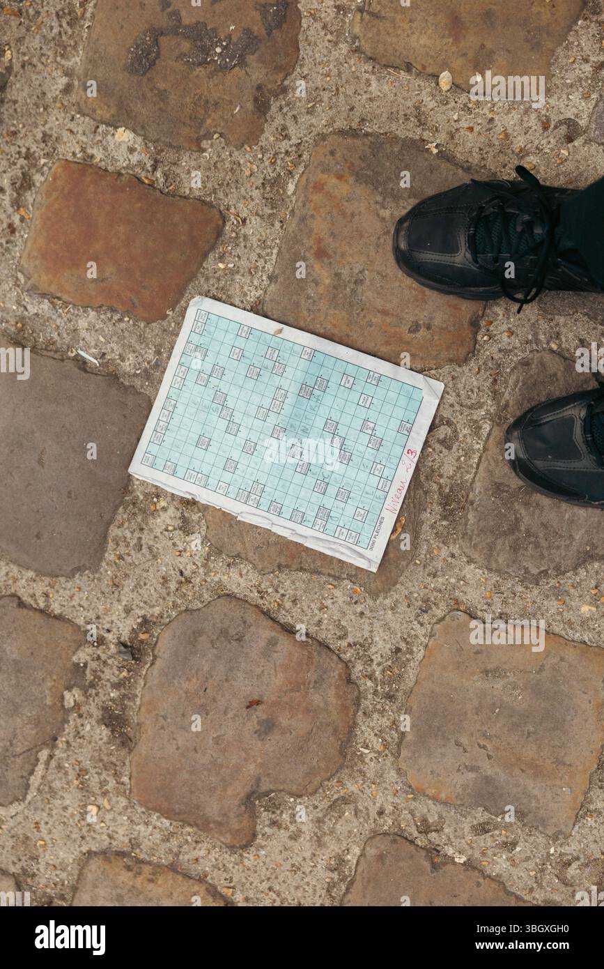 Une vue de haut en bas montre un puzzle de mots croisés partiellement terminé reposant sur un pavé texturé, avec les pointes des chaussures sombres d'une personne visible Banque D'Images