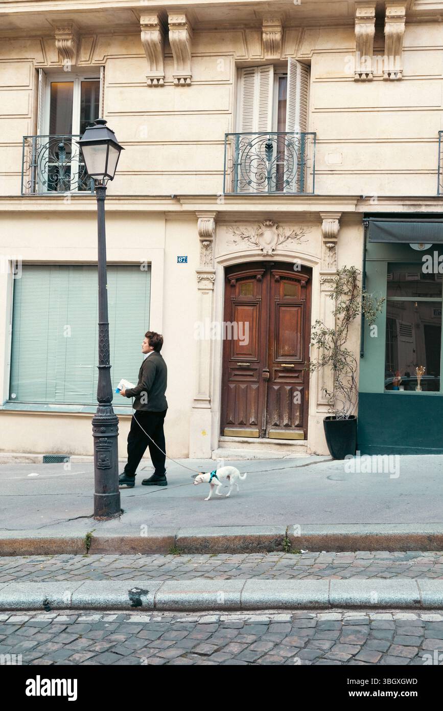 Un homme promène son petit chien blanc en laisse le long d'un trottoir pavé parisien, en passant devant un lampadaire classique, avec une façade de bâtiment traditionnelle Banque D'Images