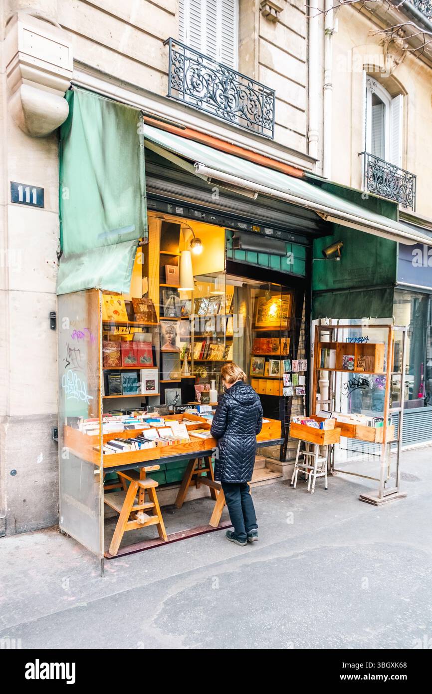 Une personne vue de dos, vêtue d'une veste matelassée, parcourt les livres exposés sur un stand extérieur devant une charmante librairie bien éclairée. Th Banque D'Images