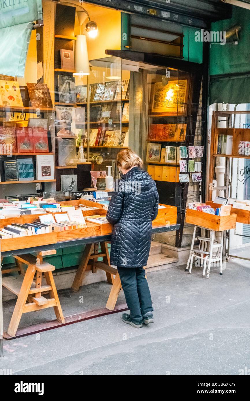 Une femme portant une veste matelassée foncée est vue de derrière, parcourez attentivement une sélection de livres disposés sur une table exposée à l'extérieur d'une librairie. Le s Banque D'Images
