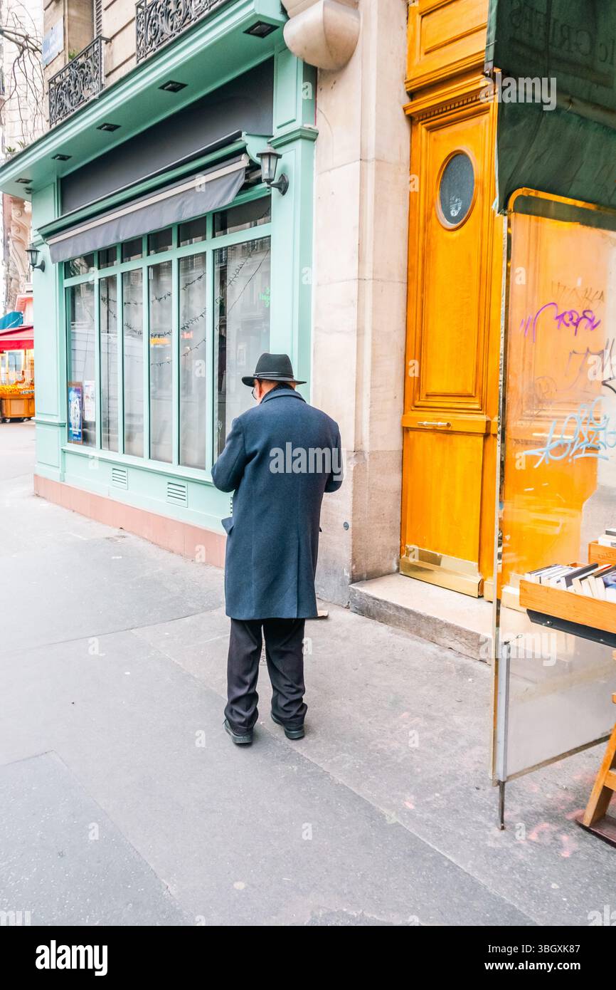 Un homme portant un pardessus sombre et un chapeau de style Fedora se tient sur un trottoir de la ville, vu de derrière. Il semble regarder vers un esprit de magasinage Banque D'Images