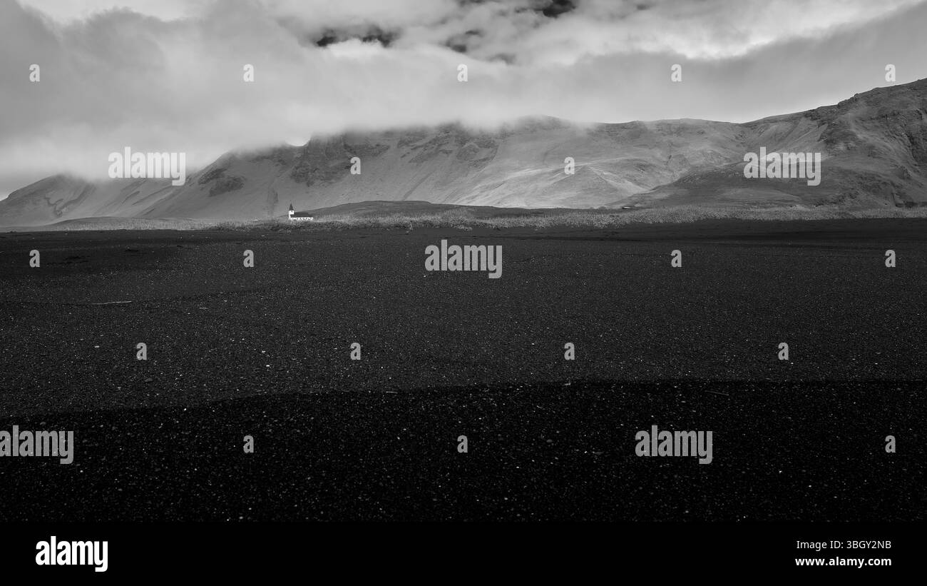 Une église isolée à Vik, en Islande, se trouve au pied de montagnes couvertes de brume. Capturé en monochrome avec un vaste sable volcanique noir au premier plan. Banque D'Images