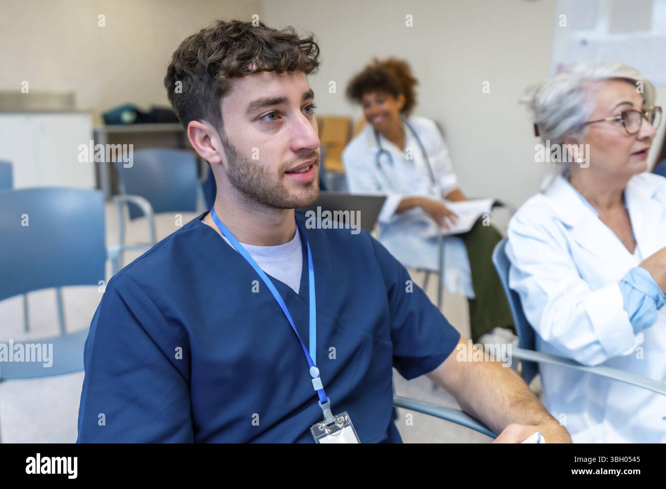 Jeune infirmier concentré à l'écoute attentive lors d'une conférence médicale, entouré de collègues en milieu hospitalier Banque D'Images