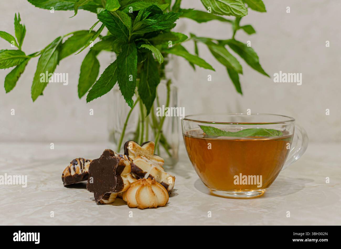 Une tasse en verre transparent remplie de tisane à la menthe garnie de feuilles de menthe fraîche. En arrière-plan est un vase avec des feuilles de menthe verte fraîche Banque D'Images