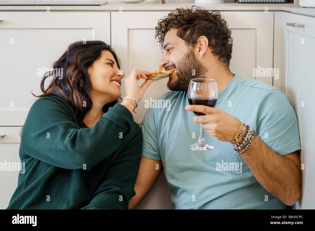Un jeune couple affectueux aime un repas décontracté et romantique à la maison, assis sur le sol de la cuisine. La femme nourrit ludique son partenaire d'une tranche de pi Banque D'Images