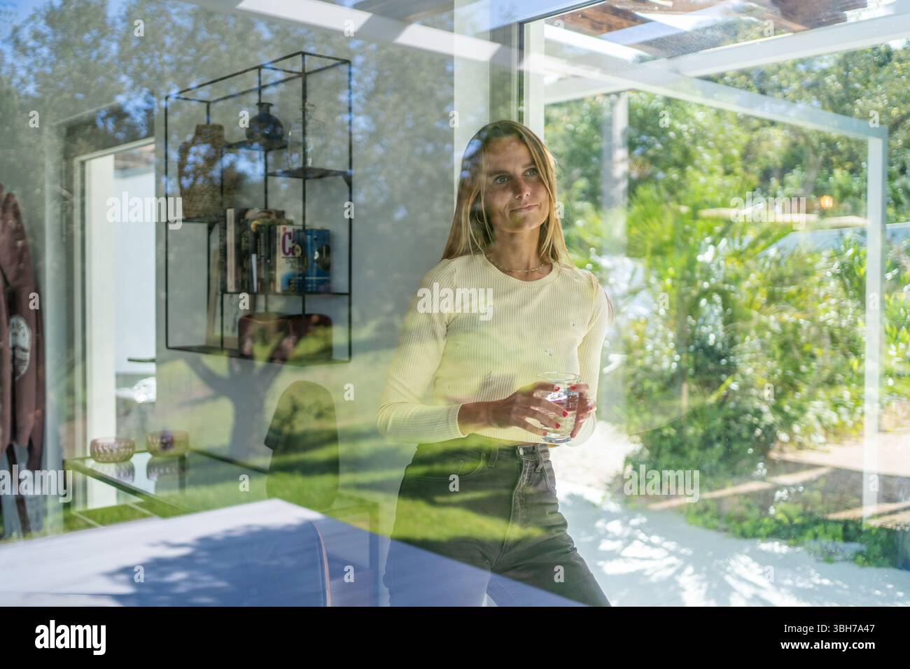 Femme, tenant un verre d'eau dans sa main. Femme regardant attentionné par la fenêtre à la maison, le matin Banque D'Images