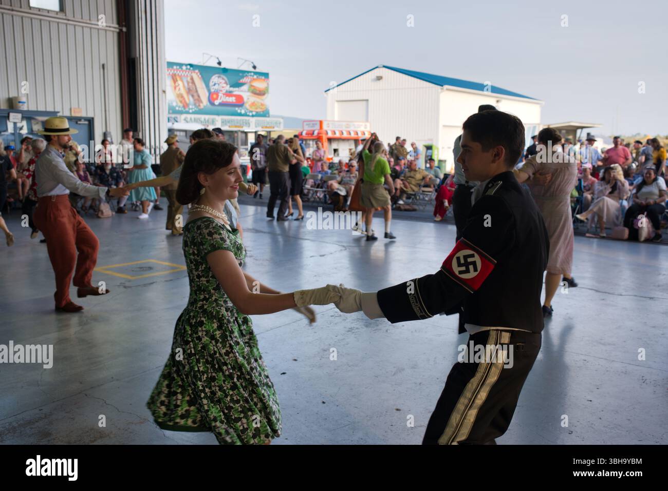 Un homme en uniforme portant une croix gammée danse avec une femme en robe verte lors d'un événement en plein air. D'autres personnes en tenue des années 1940 dansent dans le dos Banque D'Images