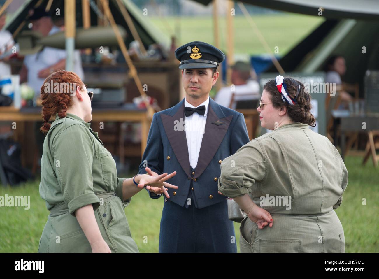 Trois individus vêtus de vêtements de l'époque de la seconde Guerre mondiale interagissent à l'extérieur. Deux femmes en combinaisons vert olive conversent avec un homme en uniforme de la Royal Air Force, w Banque D'Images