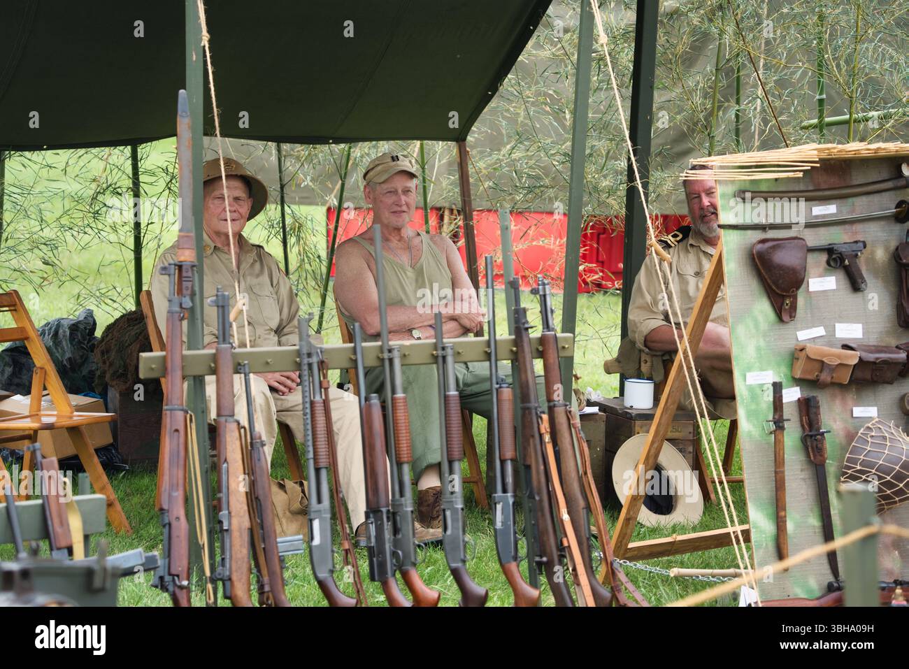 Trois hommes en tenue militaire vintage sont assis sous une tente en toile, exposant un éventail de fusils d'épaule et de souvenirs militaires sur un rack et un tableau à proximité. T Banque D'Images