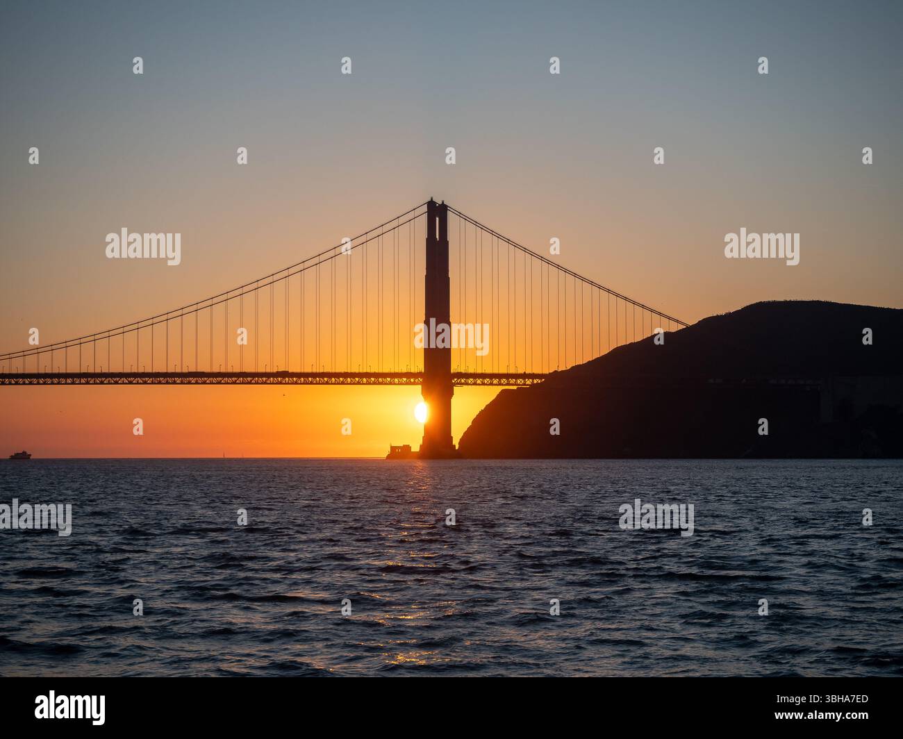 Le soleil se couche derrière le Golden Gate Bridge, créant une silhouette du monument emblématique sur le ciel coloré. L'eau reflète les teintes chaudes du coucher de soleil, ajoutant à la beauté de la scène. Banque D'Images
