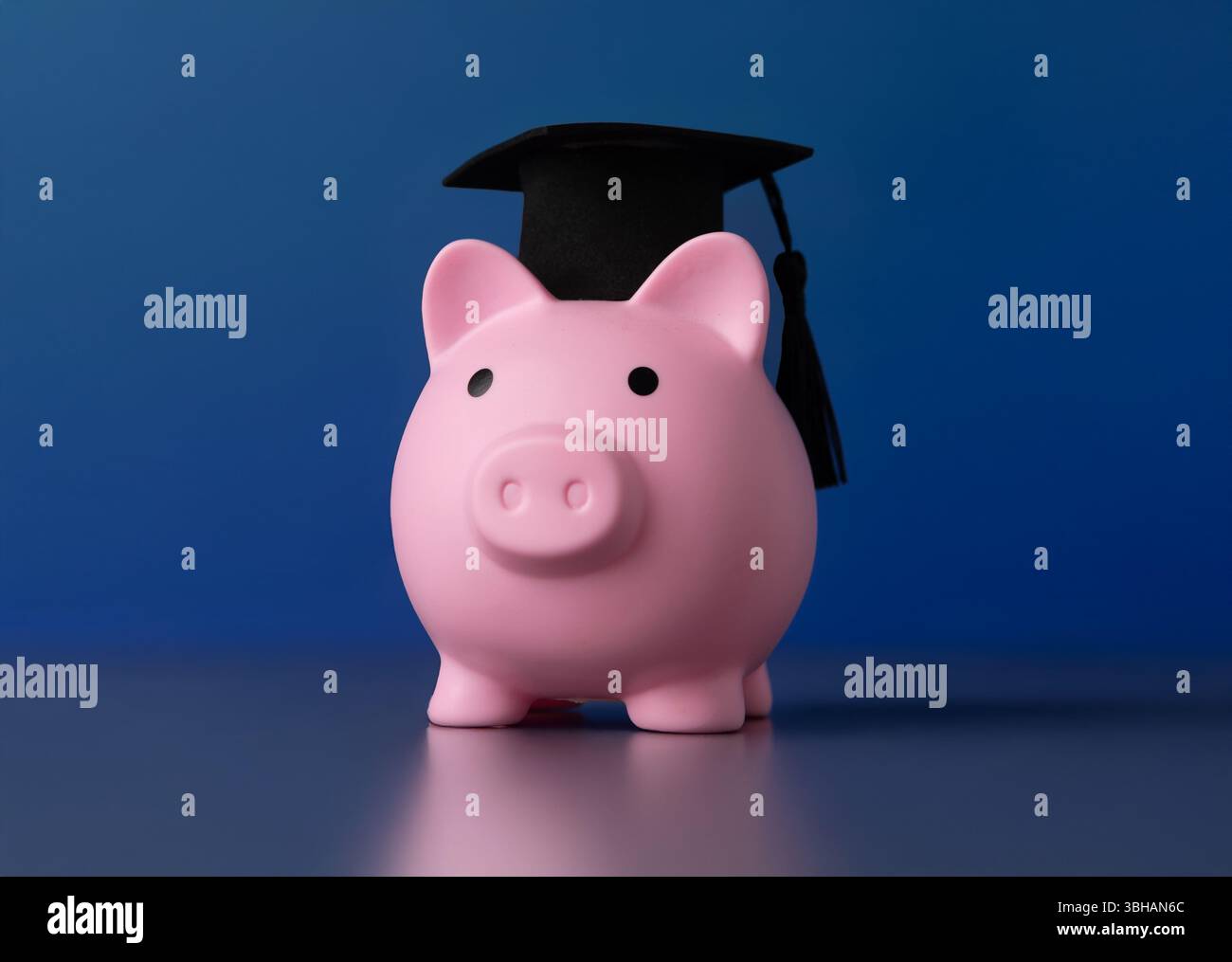 Une tirelire rose et une casquette de graduation sur un fond bleu foncé solide. Concept d'éducation, d'épargne ou d'objectifs futurs. Banque D'Images