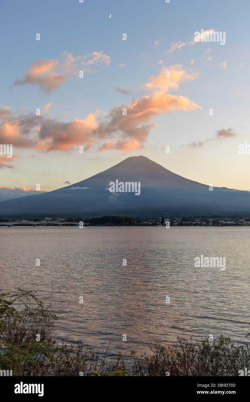 Les teintes dorées du coucher de soleil mettent en valeur les pentes du mont Fuji alors que des nuages flottent au-dessus, tandis que le lac Kawaguchi scintille de reflets ondulants reflétant la lumière du jour qui s'estompe. Banque D'Images