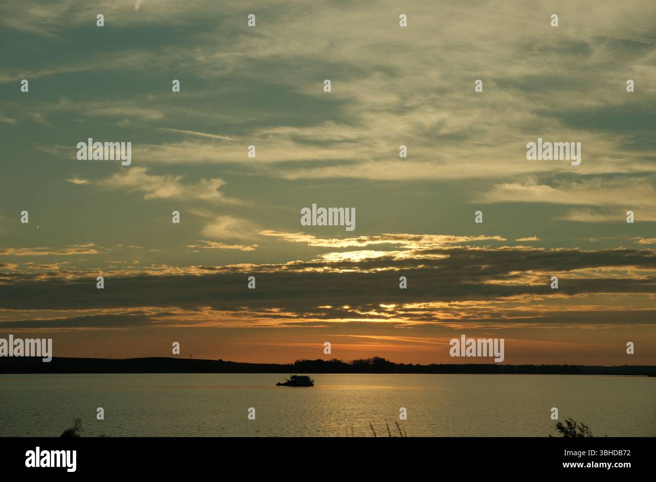 Un bateau solitaire flotte sur un lac tranquille pendant un coucher de soleil sombre. Des couches de nuages sombres et dorés s'étendent à travers le ciel, projetant des reflets. Banque D'Images