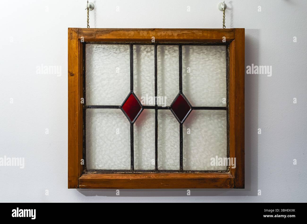 Vitrail anglais encadré en bois avec verre à motif diamant rouge suspendu par des chaînes sur un mur blanc Banque D'Images